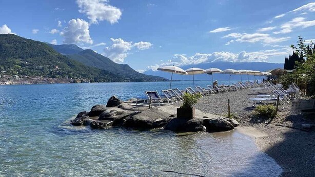 Una playa junto al lago con aguas cristalinas, orilla rocosa y sillas de descanso bajo sombrillas.