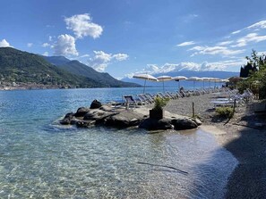 Una playa junto al lago con aguas cristalinas, orilla rocosa y sillas de descanso bajo sombrillas.