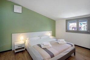 A neatly made bed with pillows and a folded towel, a bedside lamp, and a window with a view of buildings.