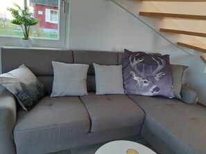 Un canapé d’angle gris avec trois coussins, une plante en pot posée sur un rebord de fenêtre et un escalier sur le côté droit.