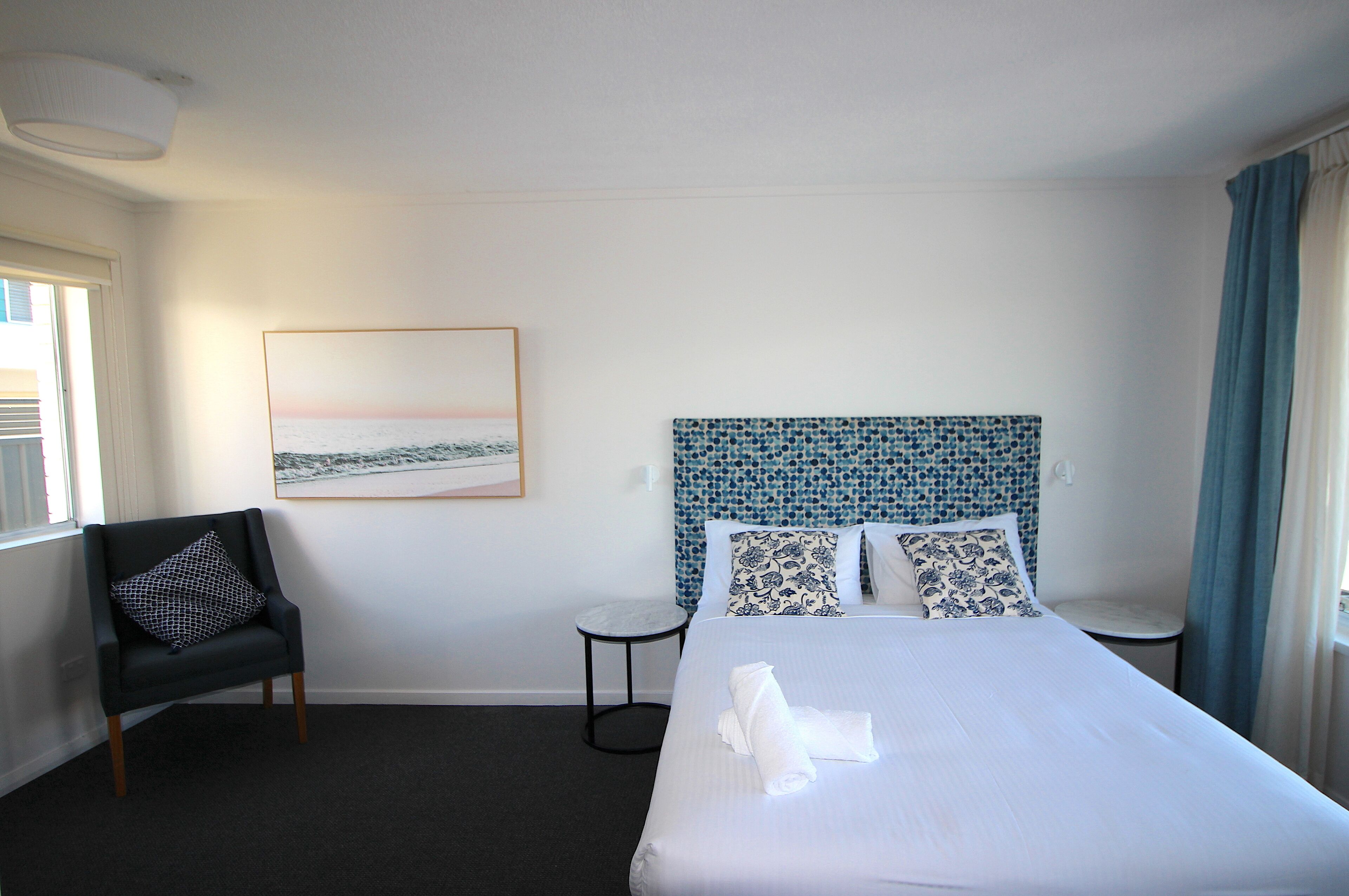 A hotel room with a large bed, a chair, a small table, and a painting on the wall.