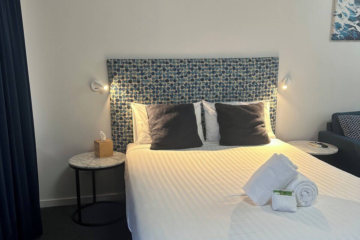 A hotel room with a bed, a headboard with a blue pattern, a bedside table with a tissue box, and a small round table with a remote control.