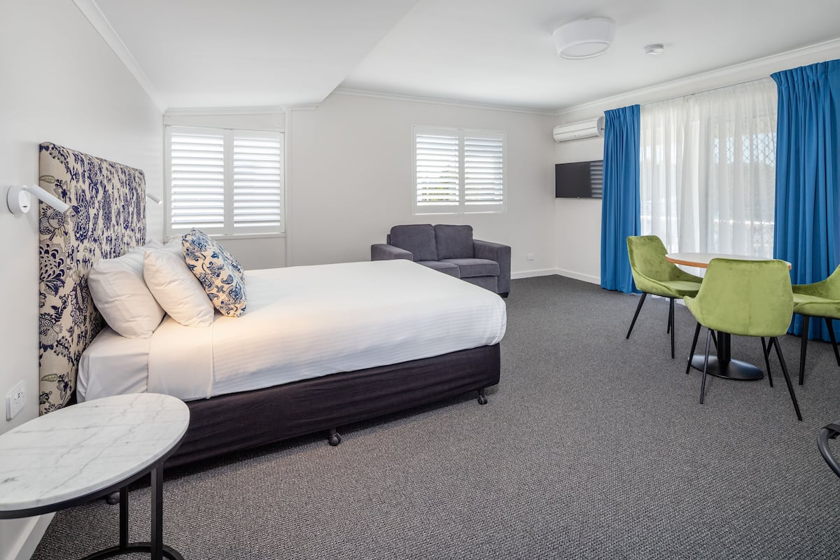 A hotel room with a large bed, a small sofa, a dining table with chairs, and a TV on the wall.