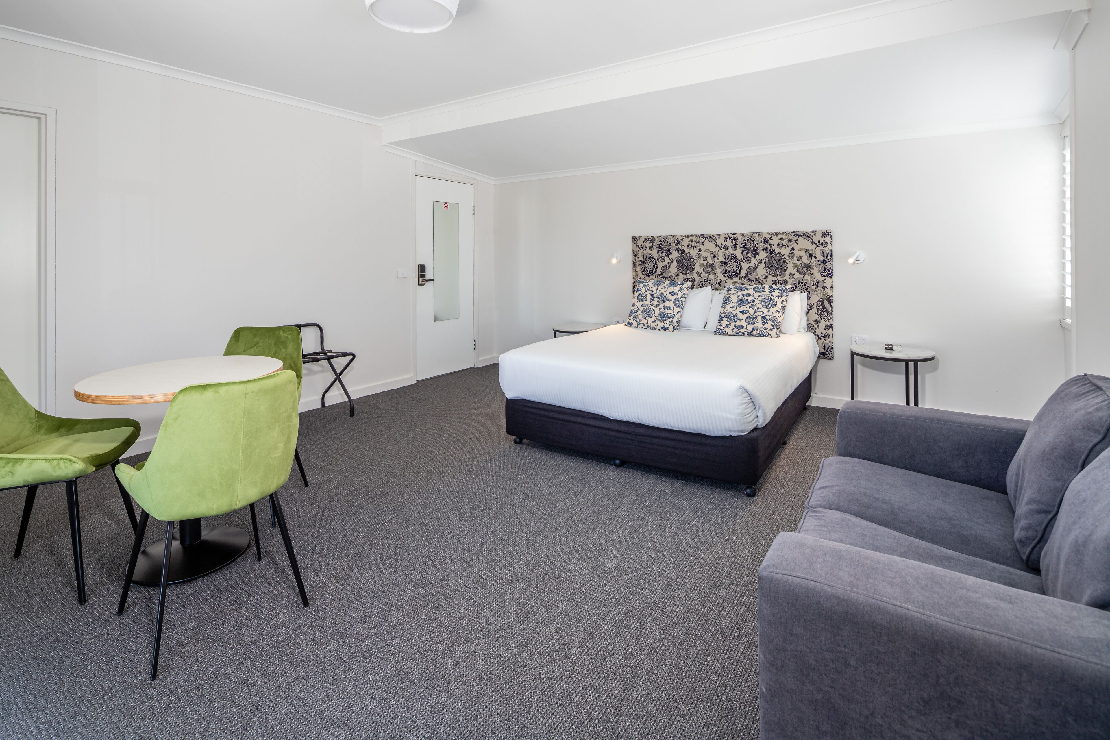Habitación de hotel con cama, sofá gris, sillón verde, una mesita redonda y una puerta blanca.