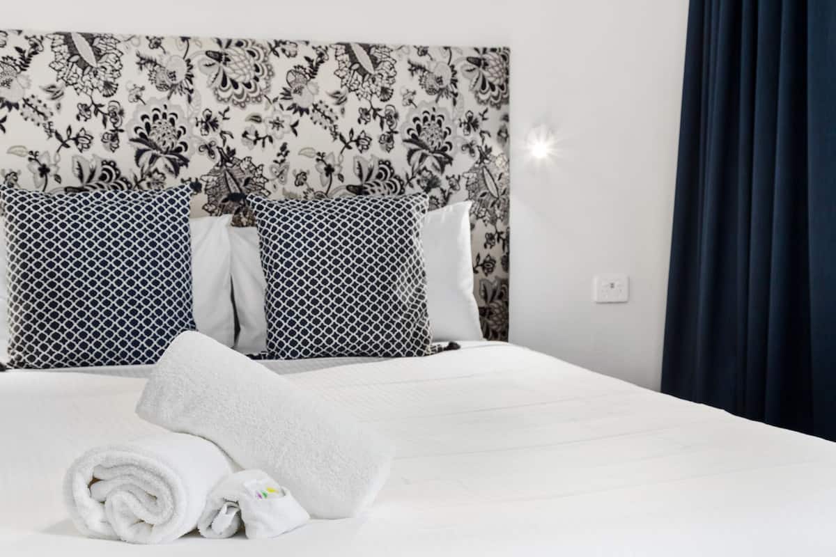 A hotel room with a bed, white linens, patterned headboard, and a dark curtain.