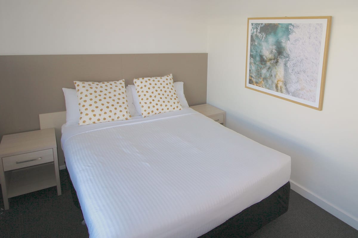 A neatly made bed with white linens and polka-dotted pillows, a small bedside table with a drawer, and a framed abstract painting on the wall.