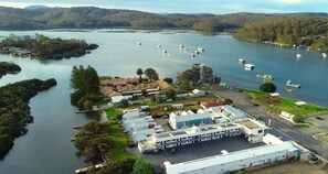An aerial view of a lakeside property with a large building, parking area, and boats on the water. - Batemans Bay Lodge (Batemans Bay)