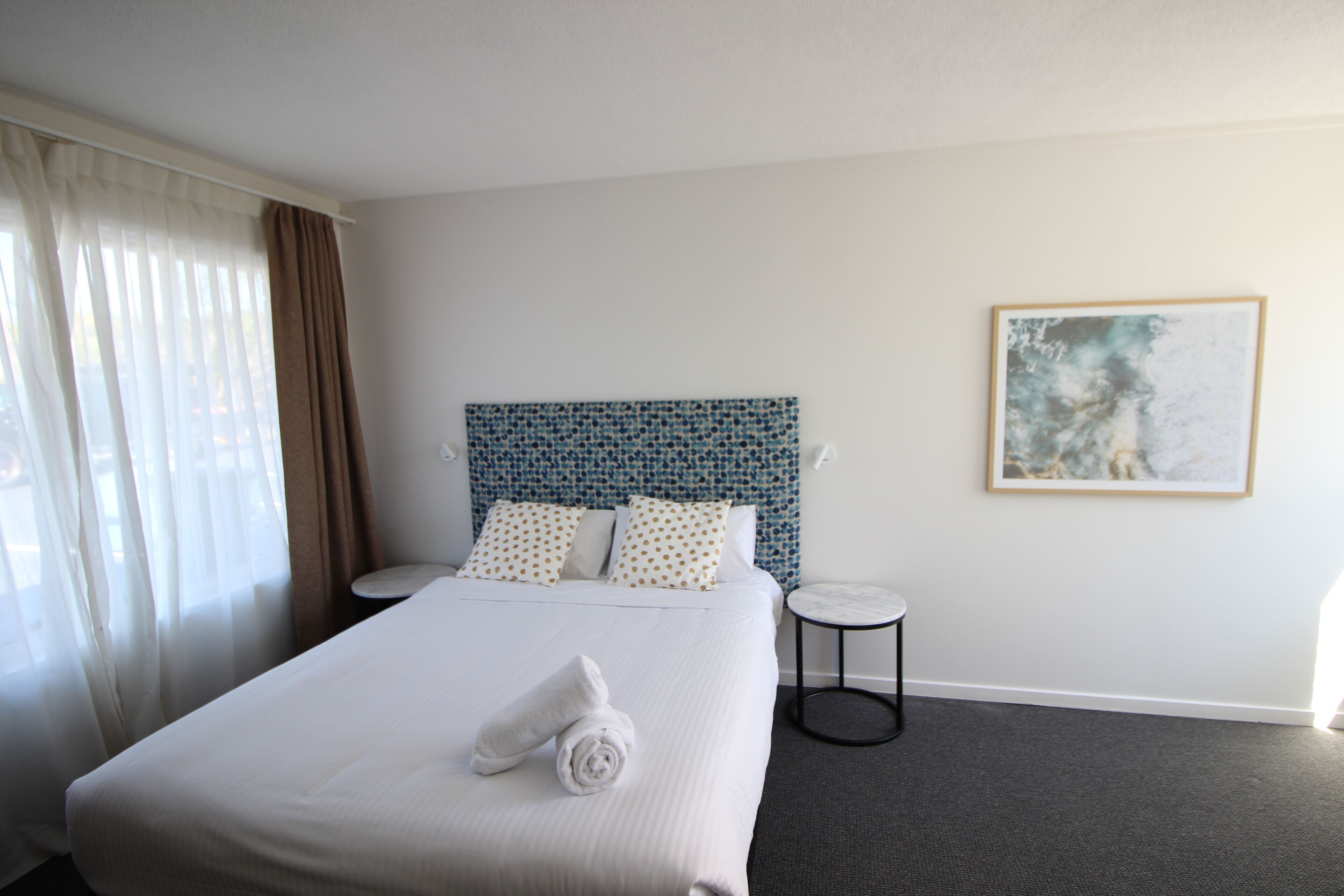 A hotel room with a large bed, a headboard with a blue pattern, a round side table, and a framed picture on the wall.