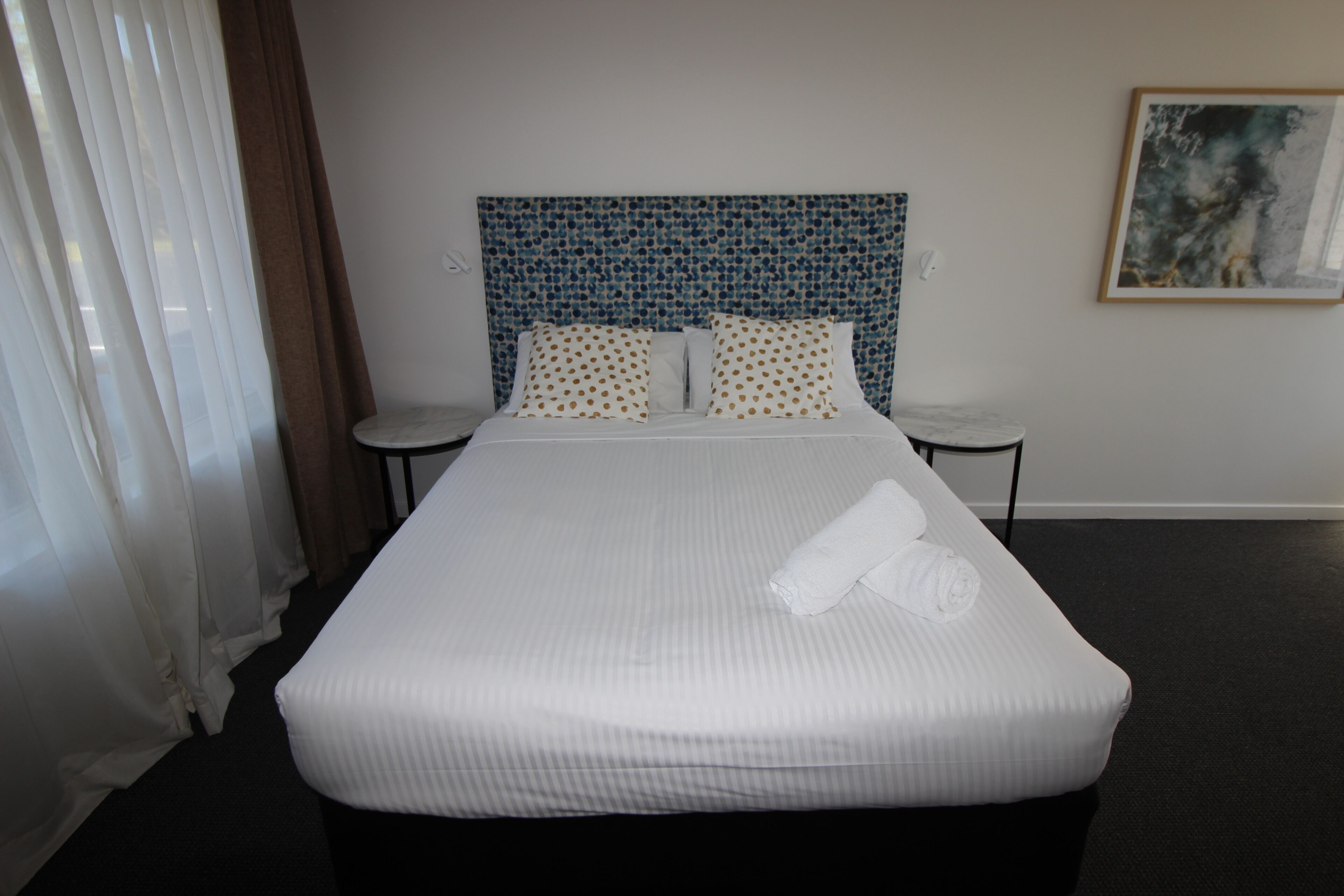 Una habitación de hotel con una cama grande, ropa de cama blanca, un cabecero con estampado azul, dos mesitas de noche y un cuadro en la pared.