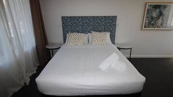 Una habitación de hotel con una cama grande, ropa de cama blanca, un cabecero con estampado azul, dos mesitas de noche y un cuadro en la pared.