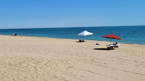 A beach with a white tent and a red umbrella, a lounge chair, and a few people in the distance.