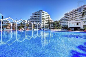 Une piscine à l’eau d’un bleu limpide, entourée de bâtiments blancs dotés de balcons, avec un palmier au premier plan.