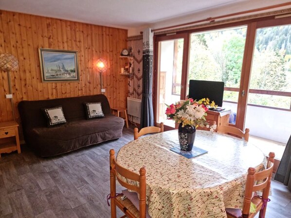 A cozy room with a wooden wall, a sofa, a dining table with chairs, a vase of flowers, and a television.