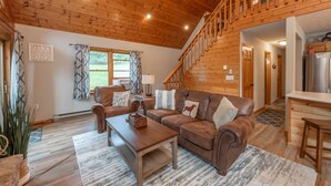 A cabin interior with a wooden ceiling, a brown leather sofa, a wooden coffee table, and a staircase leading to an upper level. - Dog Friendly Cozy Escape w/ Hot Tub & Porch Views (Swanton)