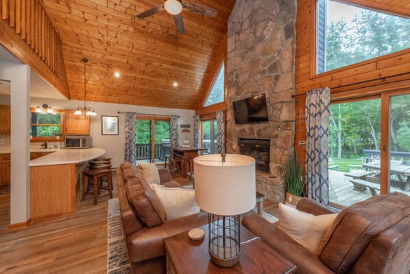 A spacious living area with a stone fireplace, wooden ceiling, and large windows overlooking a deck.