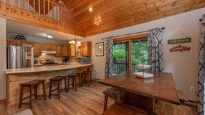 A wooden cabin interior with a kitchen, dining area, and a balcony overlooking greenery. - Dog Friendly Cozy Escape w/ Hot Tub & Porch Views (Swanton)