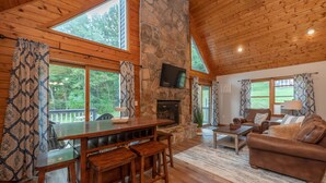 A cozy cabin interior with a stone fireplace, wooden furniture, and large windows offering a view of the forest. - Dog Friendly Cozy Escape w/ Hot Tub & Porch Views (Swanton)
