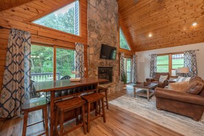 A cozy cabin interior with a stone fireplace, wooden furniture, and large windows offering a view of the forest.