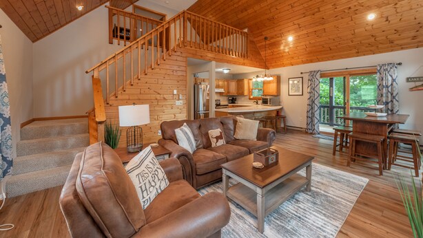 A spacious living area with a wooden staircase, a sofa set, a dining table, and a kitchen in the background. - Dog Friendly Cozy Escape w/ Hot Tub & Porch Views (Swanton)