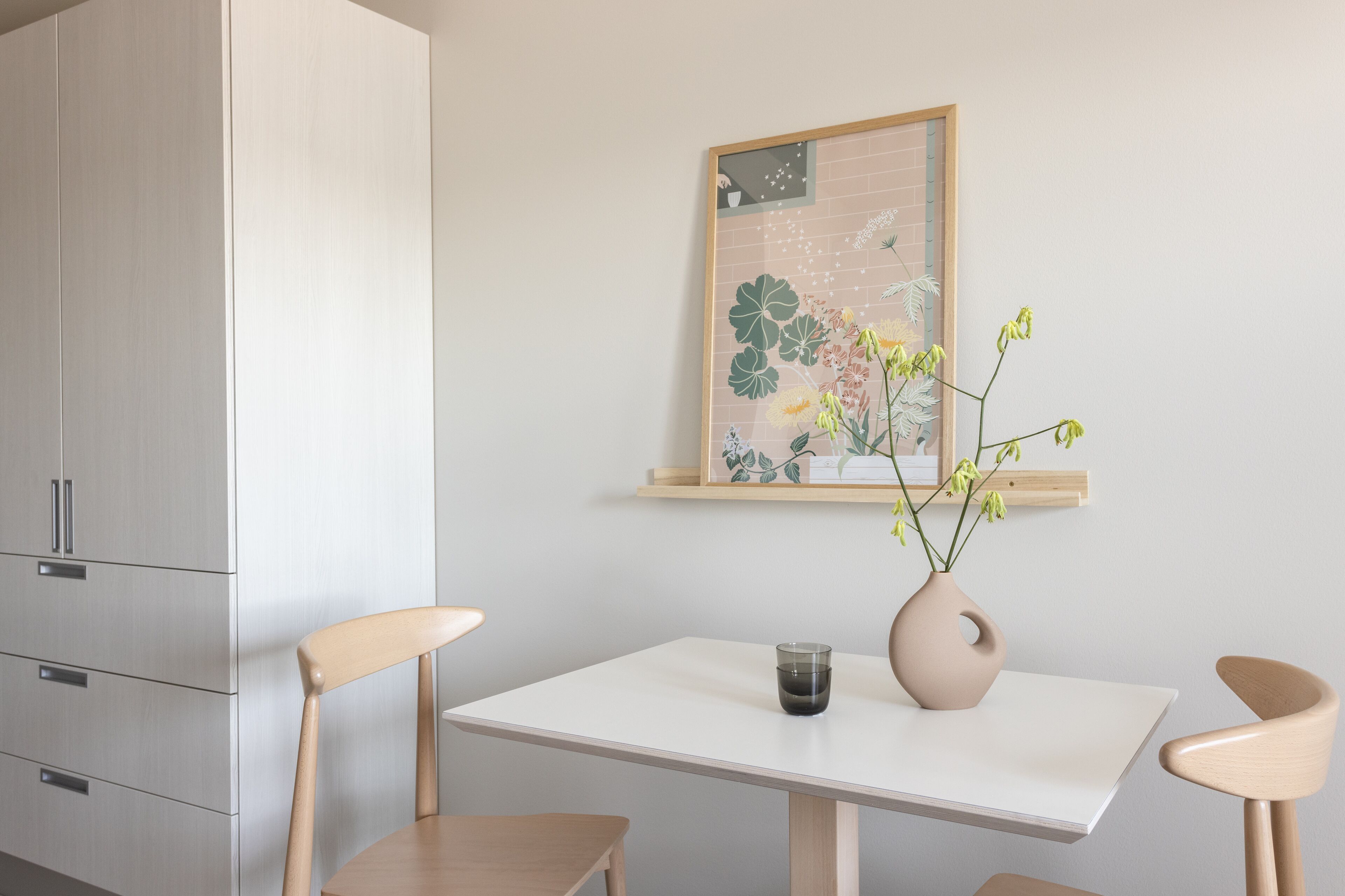 Un comedor minimalista con una mesa blanca, dos sillas de madera, un jarrón con flores y una obra de arte enmarcada en la pared.