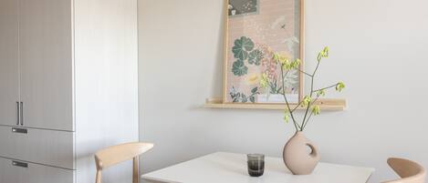 A minimalist dining area with a white table, two wooden chairs, a vase with flowers, and a framed artwork on the wall.