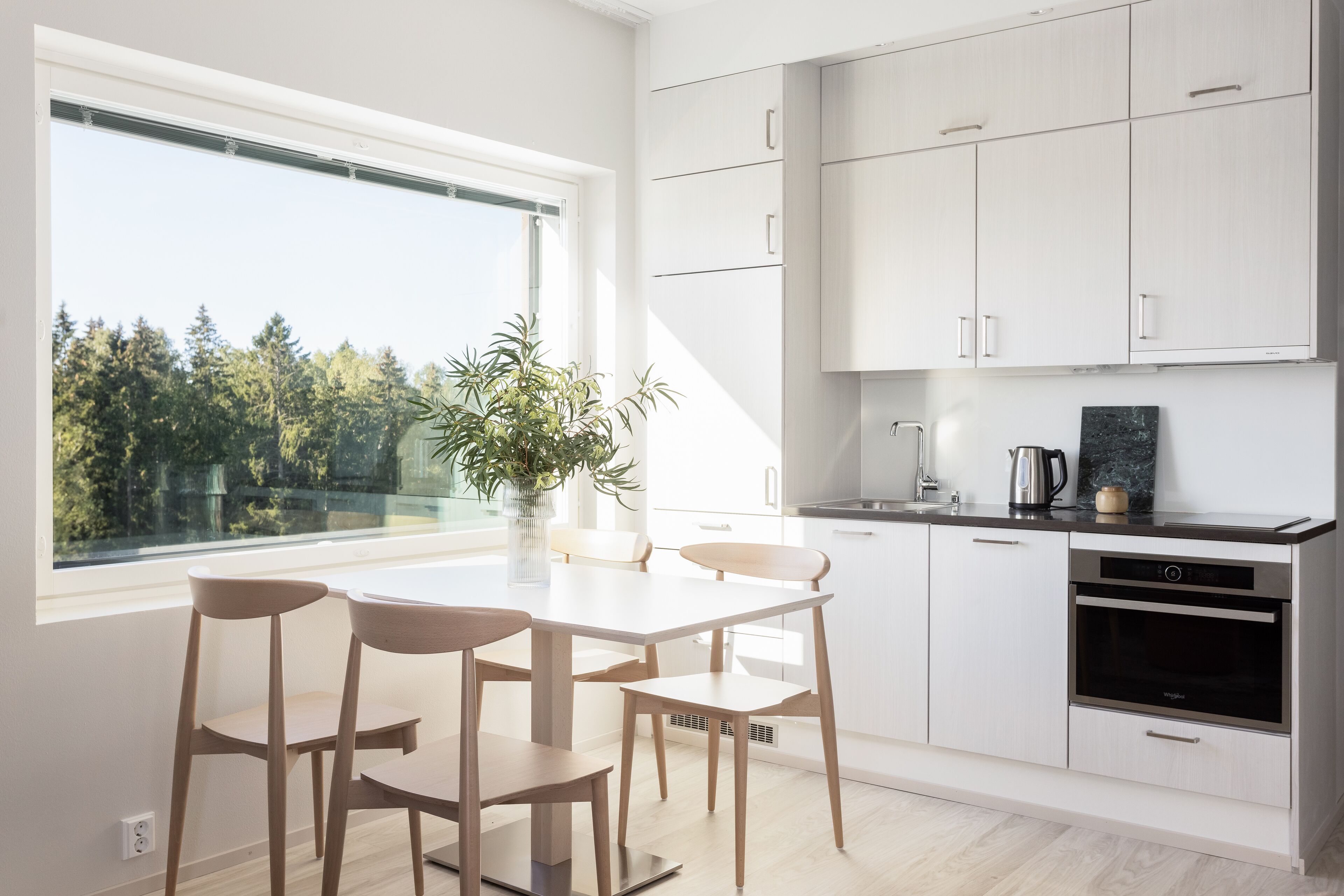 A modern kitchen with white cabinets, a dining table with chairs, a window with a view of trees, and a coffee maker.