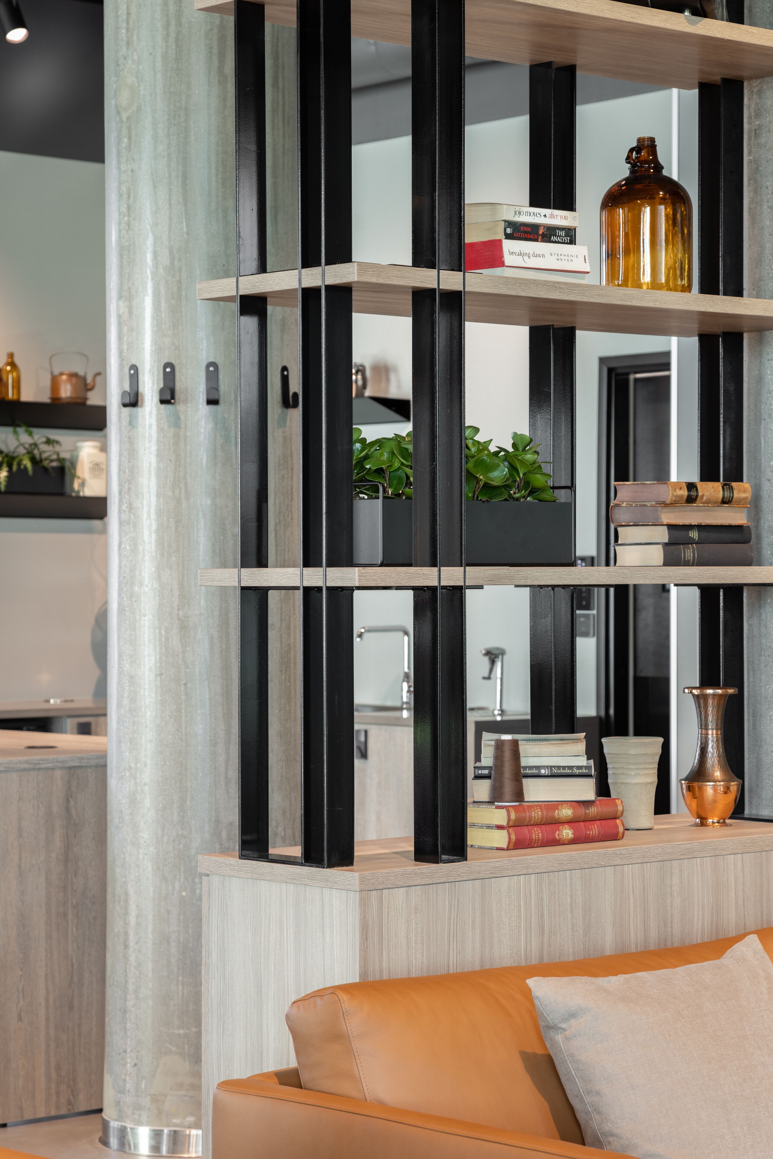 Una sala de estar moderna con un sofá, una estantería llena de libros y objetos decorativos, y una cocina al fondo.