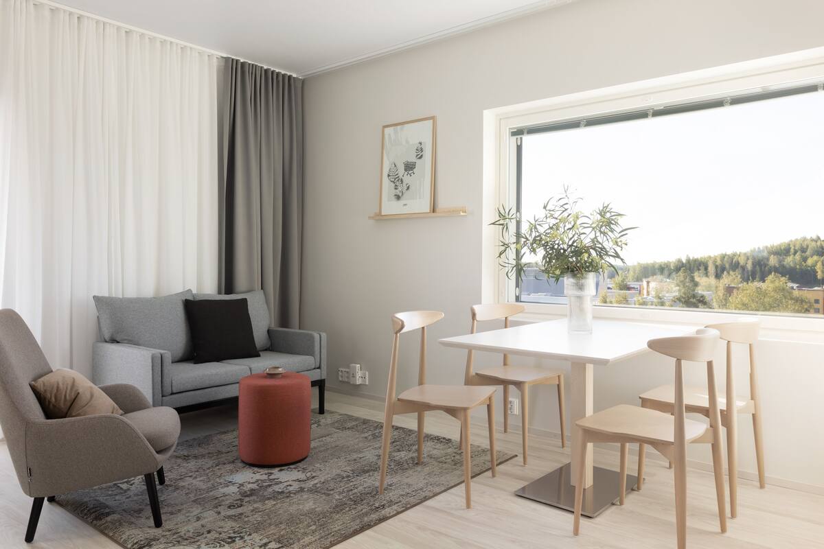 Ein modernes Wohnzimmer mit einem grauen Sofa, einem roten Hocker und einem Esstisch mit vier Stühlen. Ein großes Fenster bietet Blick auf Bäume und Gebäude.