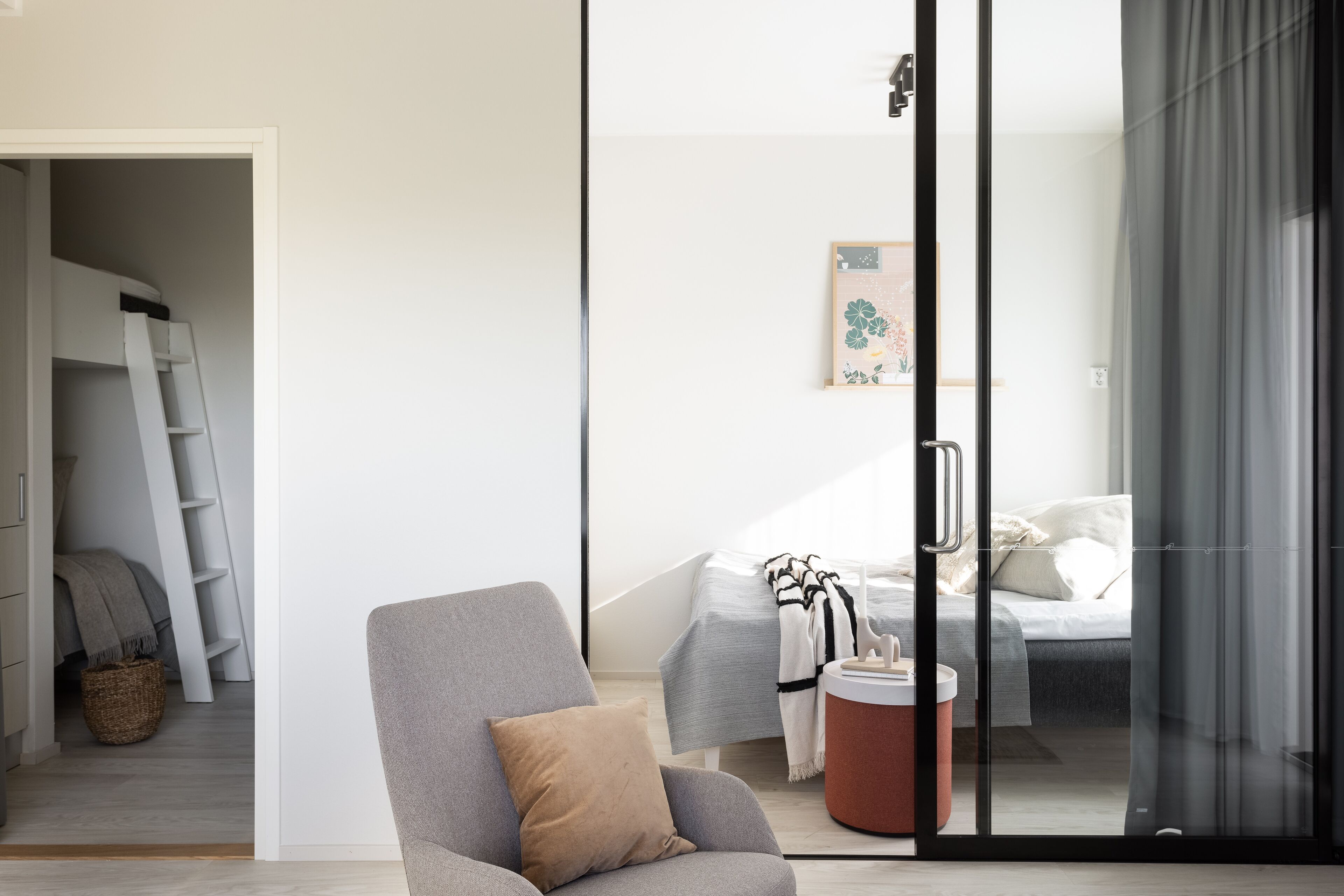 Un dormitorio moderno con puerta de vidrio, una cama con manta a rayas, un sillón gris y una pequeña mesa redonda.