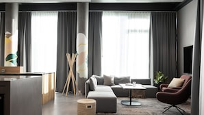 Una sala de estar moderna con un sofá gris, un sillón rojo y una pequeña mesa de centro. Hay un ventanal con cortinas grises, un televisor de pantalla plana y una mesita de noche de madera.
