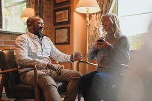 Dos personas sentadas en una habitación con pared de ladrillo, una lámpara y cuadros enmarcados.