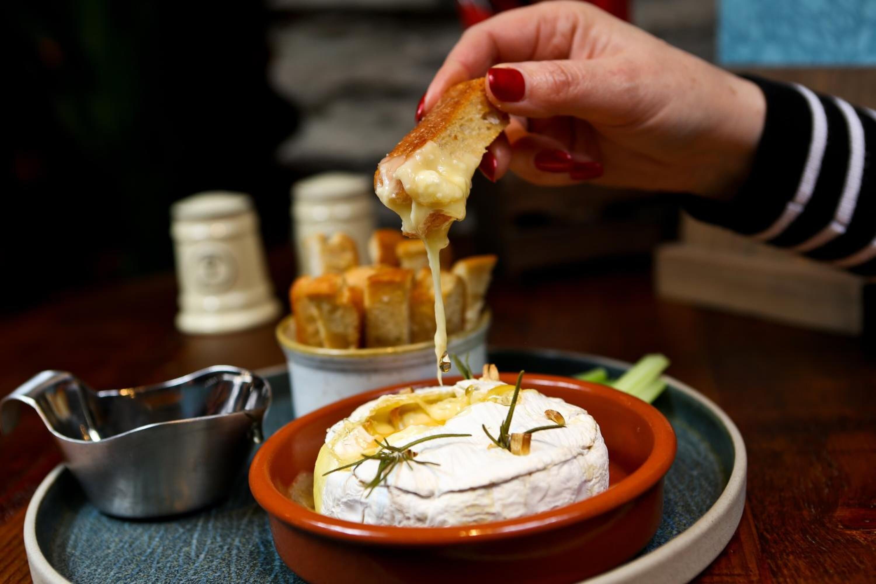 A hand holding a piece of bread with melted cheese, a bowl of cheese with herbs, and a small metal container on a table.
