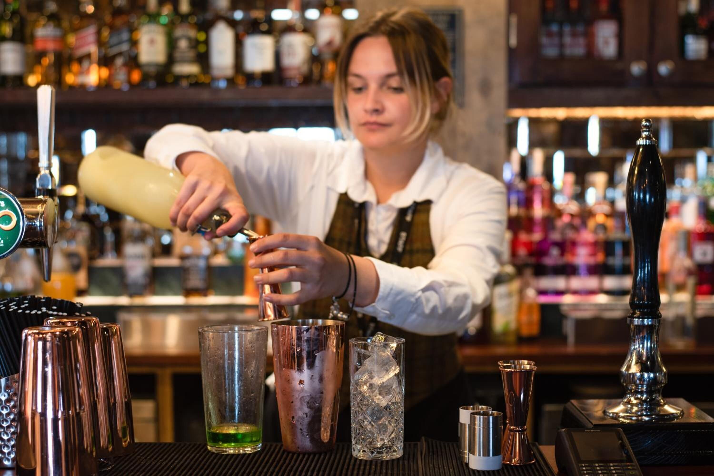 A bartender preparing a drink at a bar with various bottles and bar tools in the background.