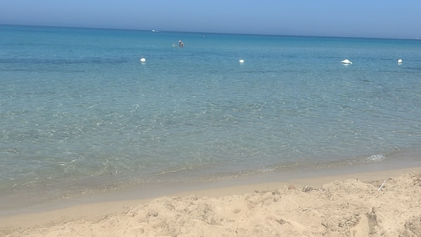 Una platja amb aigua cristal·lina, un tram de sorra i alguns boies a la distància.