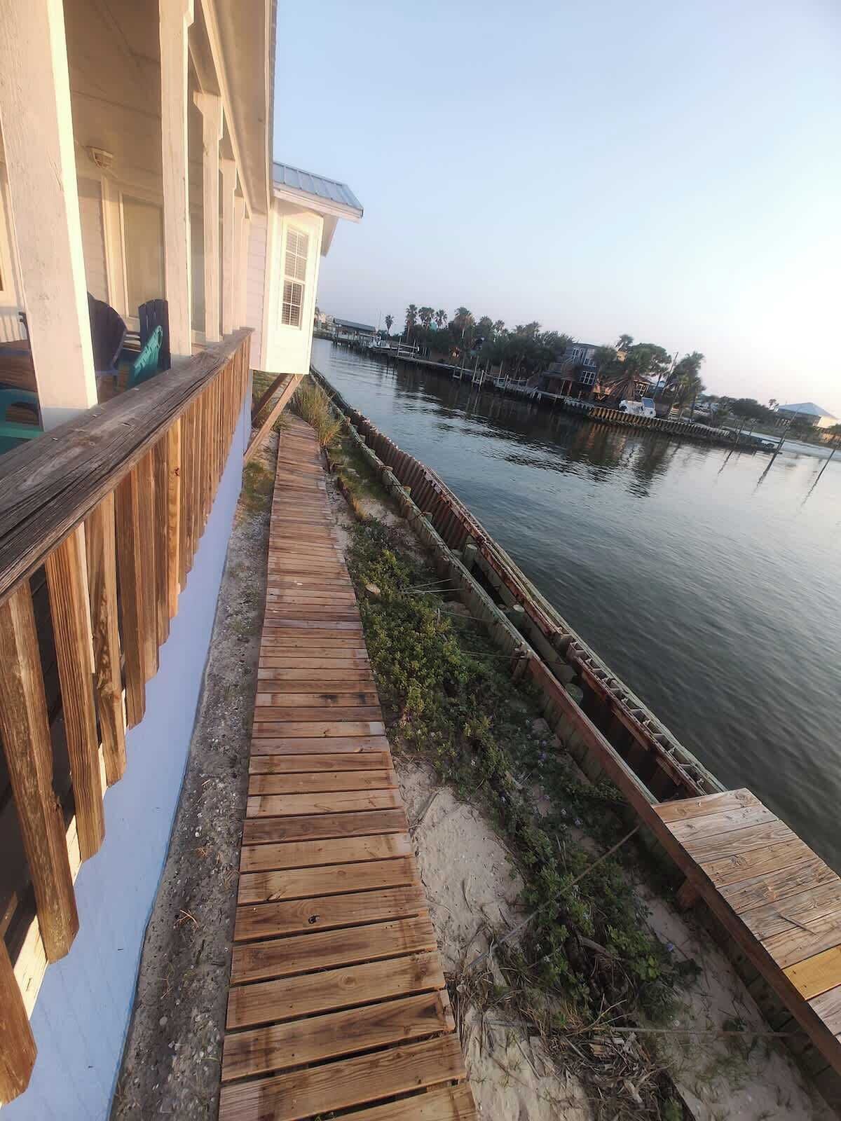 Eine Holzterrasse neben einem Gewässer, mit einem Gebäude an einer Seite und einem Steg an der anderen.