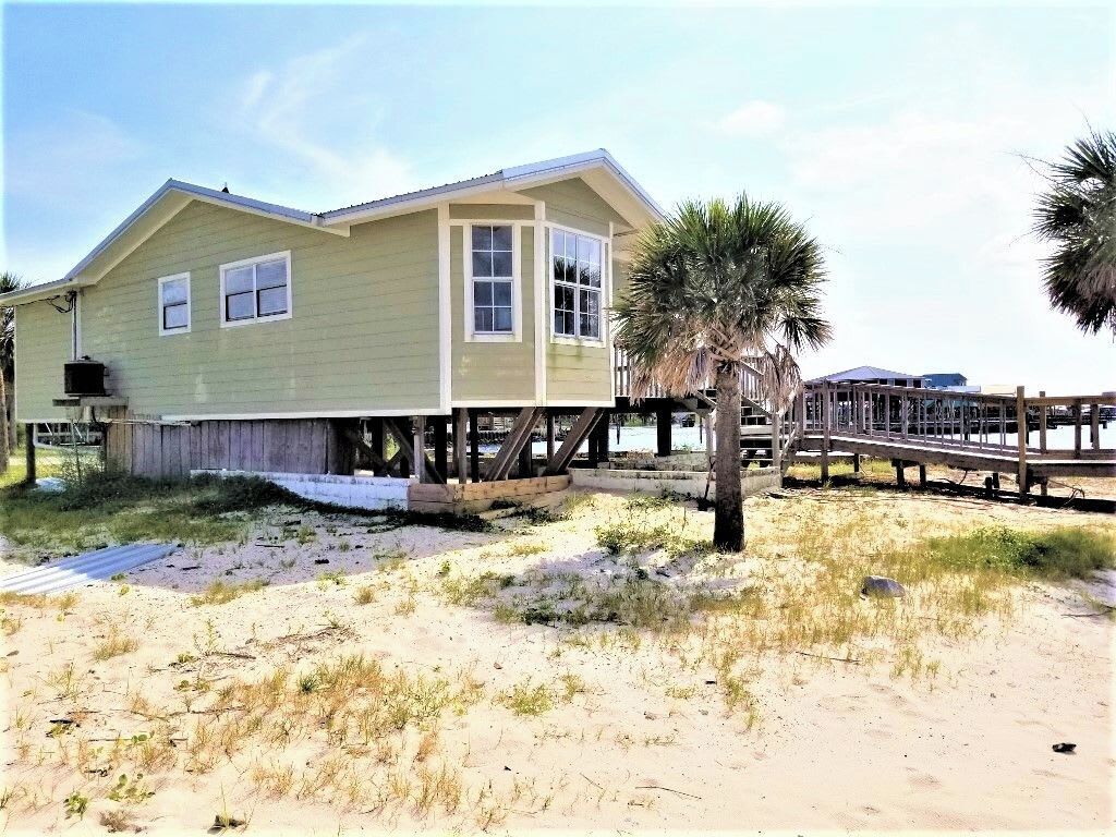 Una casa de playa con terraza de madera, una palmera y un muelle que se adentra en el agua.
