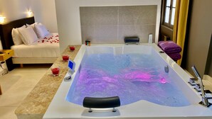 A modern bathroom with a large bathtub, a bedside table, and a bed with white bedding.