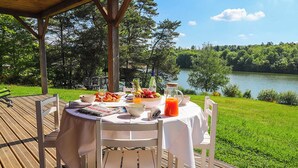 An outdoor dining area with a table set for two, surrounded by a wooden deck and overlooking a lake.