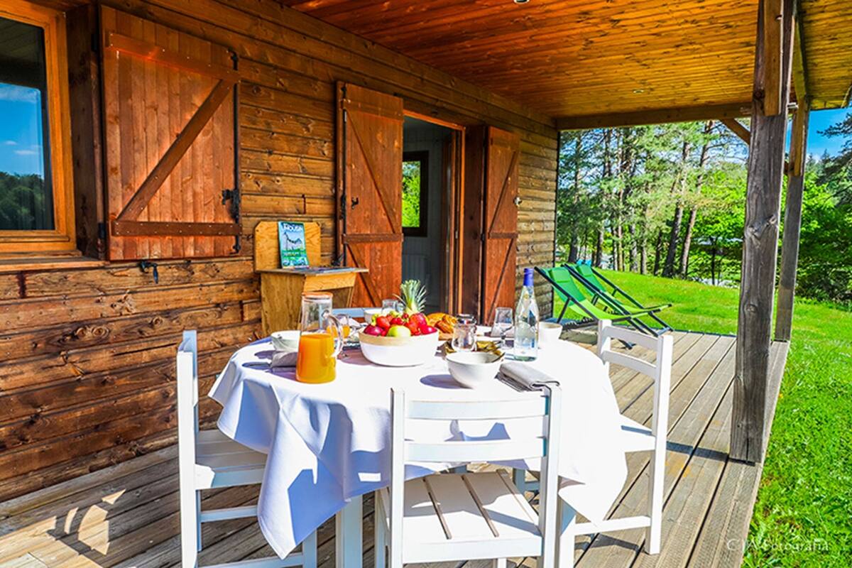 Una cabaña de madera con una mesa lista para el desayuno, un césped verde y una vista despejada del bosque circundante.