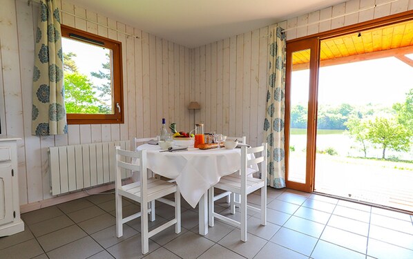 Un coin repas avec une table dressée pour quatre personnes, des chaises blanches, un radiateur, une fenêtre avec des rideaux et une vue sur la verdure à l’extérieur.