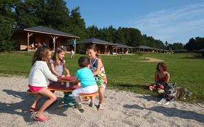 Niños jugando en juegos infantiles al aire libre, un perro y un edificio con un porche al fondo.