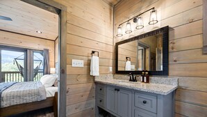 A wooden cabin interior with a bed, a bathroom with a large mirror, and a window with a view of the outdoors. - BRADN NEW Luxury Cabin! The ELM/PanoramicMountainView/Modern/LARGE DECKs (Gatlinburg)