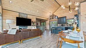 A modern living room with a brown leather sofa, a dining table with chairs, a TV mounted on the wall, and a kitchen area with a bar. - BRADN NEW Luxury Cabin! The ELM/PanoramicMountainView/Modern/LARGE DECKs (Gatlinburg)
