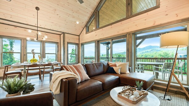 A spacious living area with a large brown leather sofa, a dining table with chairs, and a view of a wooden deck and mountains through large windows. - BRADN NEW Luxury Cabin! The ELM/PanoramicMountainView/Modern/LARGE DECKs (Gatlinburg)