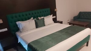 Een net opgemaakt bed met een groen hoofdeinde en witte beddengoed, een klein nachtkastje met een lamp, en een groene bank op de achtergrond.
