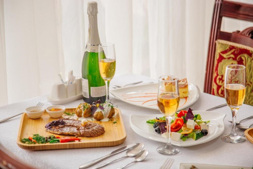 A dining table set with a bottle of white wine, glasses, a wooden board with steak and vegetables, and a plate with a salad.