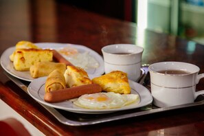 Un vassoio con prodotti per la colazione, tra cui un hot dog, uova fritte e pasticcini, accompagnato da due tazze di caffè.
