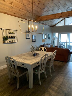 A dining area with a wooden table, white chairs, and a chandelier. There is a sofa and a window in the background.