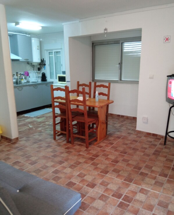 A dining area with a wooden table and chairs, a kitchen in the background, and a tiled floor.