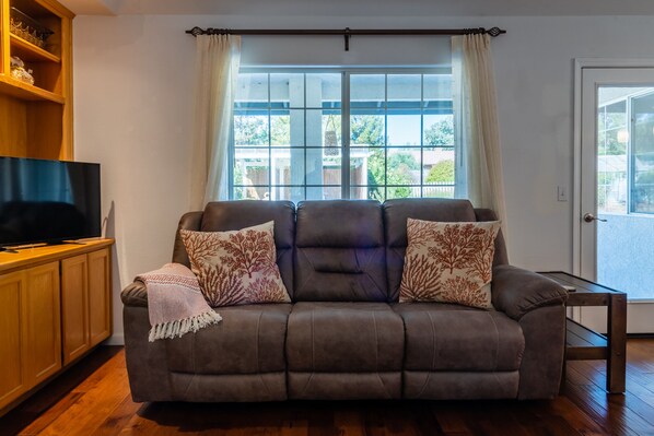 A living room with a brown sofa, two patterned throw pillows, a television on a wooden stand, and a window with curtains.